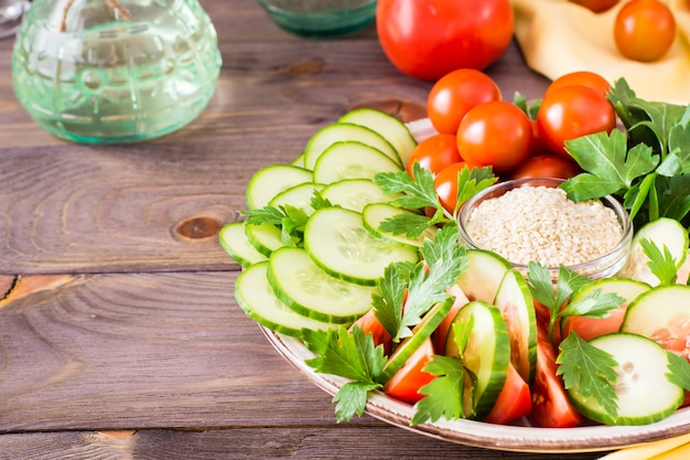 Fresh slices of cucumbers, tomatoes, sesame seeds in a bowl and parsley leaves on a plate on a wooden table