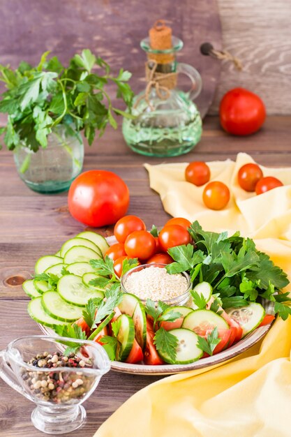 Fresh slices of cucumbers, tomatoes, sesame seeds in a bowl and parsley leaves on a plate on a wooden table