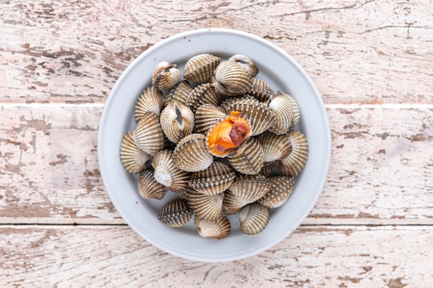 Fresh steamed cockles, boiled cockles in ceramic plate on old white wood texture background, top view, light and airy food photography, blood cockle