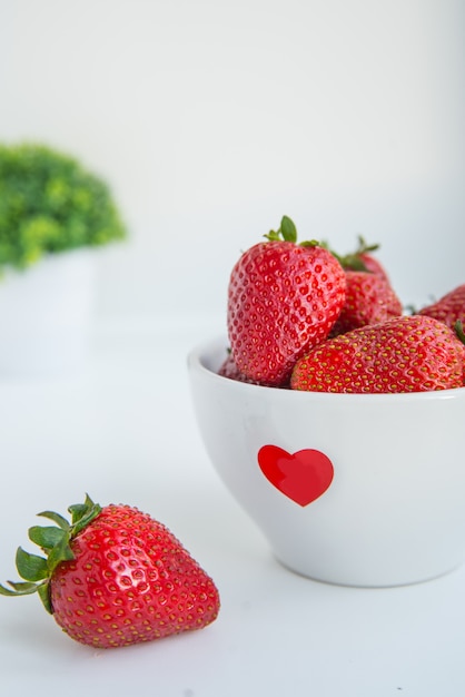 Fragole fresche in ciotola bianca con cuore rosso sui precedenti bianchi con la foto di verticale del copyspace