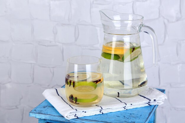 Fresh summer drink with lime and cloves in glass and jug on color wooden stand on light wall background