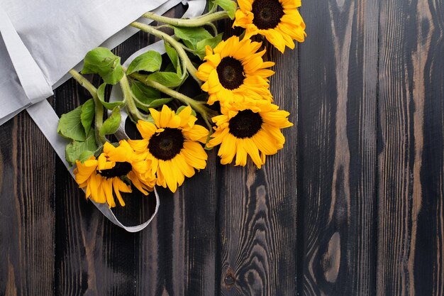 Fresh sunflowers with leaves on stalk in shopping bag on wooden background flat lay top view copy sp