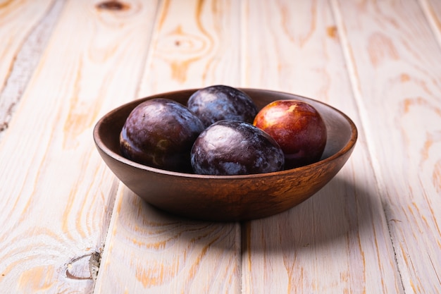 Fresh sweet plum fruits in brown wooden bowl, wood table, angle view
