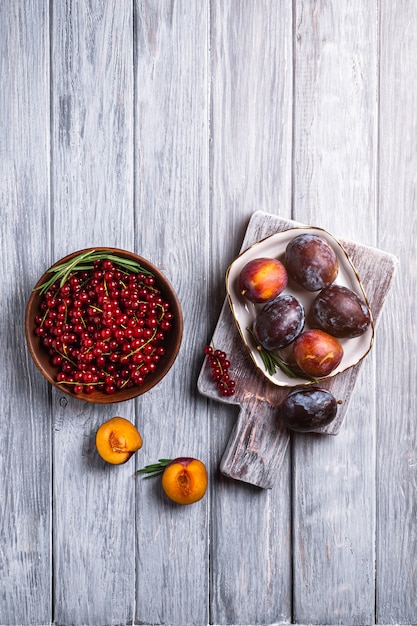 Fresh sweet plum fruits whole and sliced in plate with rosemary leaves on old cutting board with red currant berries in wooden bowl, grey wood table, top view copy space