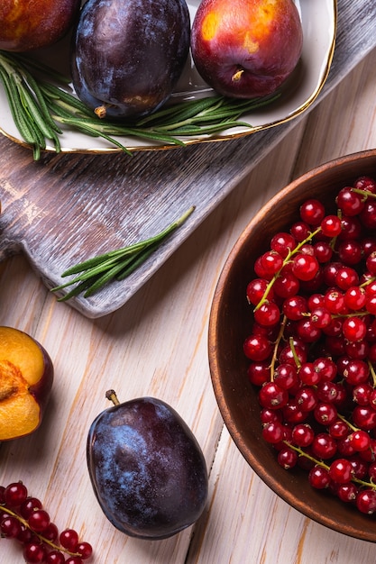 Dolce fresco prugna frutti interi e tagliati a fette nella piastra con foglie di rosmarino sul vecchio tagliere con bacche di ribes rosso nella ciotola di legno, tavolo in legno sfondo, angolo di visione