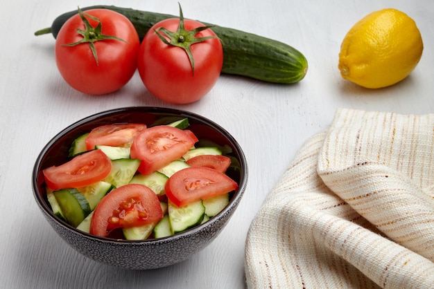 Fresh tomato and cucumber salad in dark bowl and kitchen napkin and lemon on white wooden table