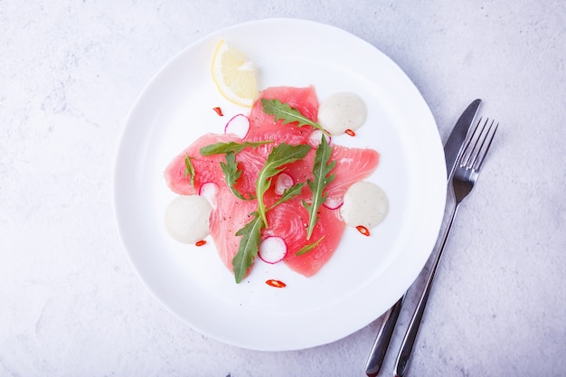 Fresh tuna carpaccio with arugula, radish, chili, lemon and sauce. Traditional cold appetizer of Italian cuisine. Close-up, selective focus, top view.