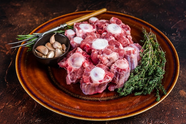 Fresh uncooked beef Oxtail cut Meat on rustic plate with herbs. Dark background. Top view.