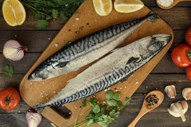 Fresh uncooked mackerel fish with lemon, herbs, oil, vegetables and spices on rustic wooden board over black backdrop, top view