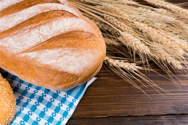 木製のテーブルの上に黄金の熟した小麦の耳を持つ新鮮なノーカットの大きな無愛想なフランスパン、クローズアップビュー