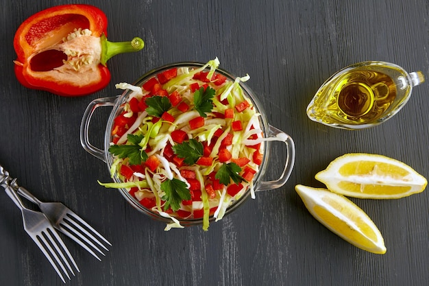 Fresh vegetable salad of cabbage and sweet peppers in a glass bowl on a black wooden background surrounded by ingredients