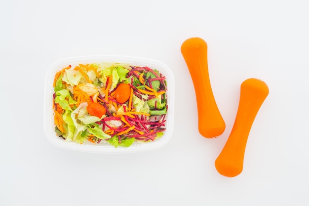 Fresh vegetable salad in lunch box with orange dumbbells on white.