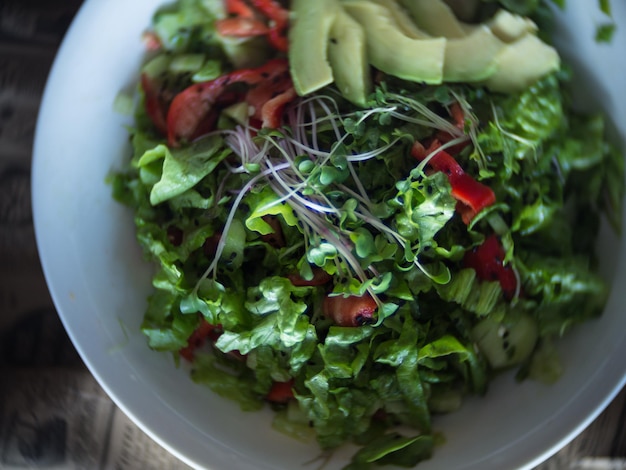 Салат из свежих овощей с огуречным салатом, авокадо, зелеными ростками, сладким перцем на белой тарелке. Миска салата с овощами на газетной абстракции.