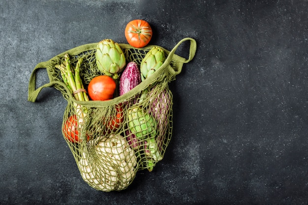 Photo fresh vegetables in a green string bag on a black table. cauliflower, tomatoes, artichokes, asparagus and zucchini.