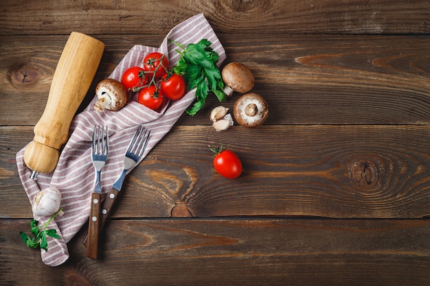 Fresh vegetables - tomatoes, garlic, champignons, parsley, pepper shaker, forks and napkin on wooden background. Place for text. Top view