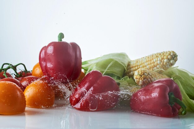 水中の新鮮な野菜が白い背景に飛び散り、トマト、ピーマン、とうもろこしが穂軸に飛び散ります。