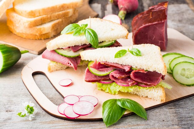 Fresh white bread sandwiches with pastrami cucumber radish and basil on a cutting board