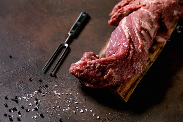 Fresh whole raw beef tenderloin meat on wooden board with metal meat fork, salt and pepper over dark brown texture background. Food cooking background concept. Close up, copy sapce