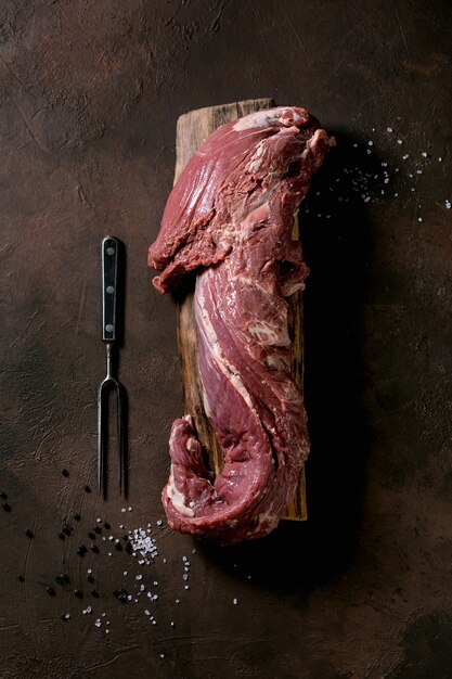 Fresh whole raw beef tenderloin meat on wooden board with metal meat fork, salt and pepper over dark brown texture background. Food cooking background concept. Top view, flat lay