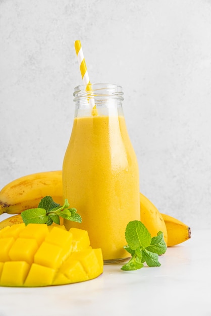 Fresh yellow mango and banana smoothie in a glass bottle with straw and mint on white background