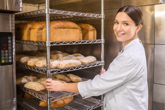 Свежеиспеченный хлеб. Красивая женщина, повернувшись лицом к камере, стоит возле духовки и подносов с хлебом в отличном настроении