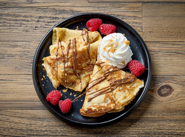 Freshly baked crepes with whipped cream and raspberries on wooden kitchen table top view