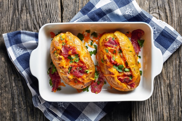 Freshly Baked hot Potatoes loaded with crispy fried Bacon, pulled chicken breast and melted cheddar cheese in a baking  dish on a rustic wooden table, view from above, close-up