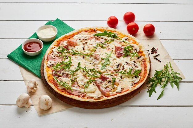 Freshly baked pizza with ham rukkola and mushrooms served on wooden background with tomatoes sauces and herbs Food delivery concept Restaurant menu