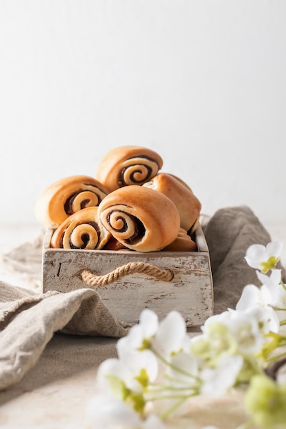 Freshly baked sweet cinnamon buns for breakfast in rustic wooden box with white flowers and linen napkin on white background Easter traditional pastry food with text space