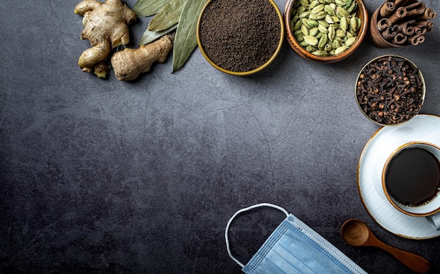 Freshly brewed tea with tea herbs and raw ingredients with face mask top view mockup dark background