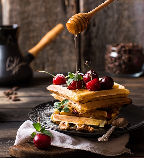 Waffle belgi appena fatti con miele e zucchero a velo. ciliegie sopra le cialde sullo scrittorio di legno e sul tovagliolo su fondo di legno. chicchi di caffè in barattolo di vetro. caffettiera turca per colazione