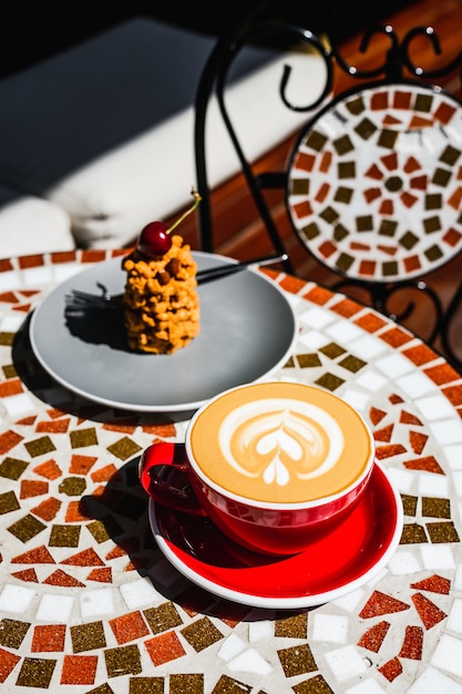 Photo a freshly made red cup of cappuccino coffee and a dessert on a mosaic stone table of a cafe terrace on a sunny day