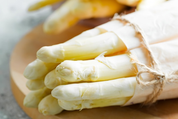 Freshly picked raw organic white asparagus bundles on wood board on light grey stone.