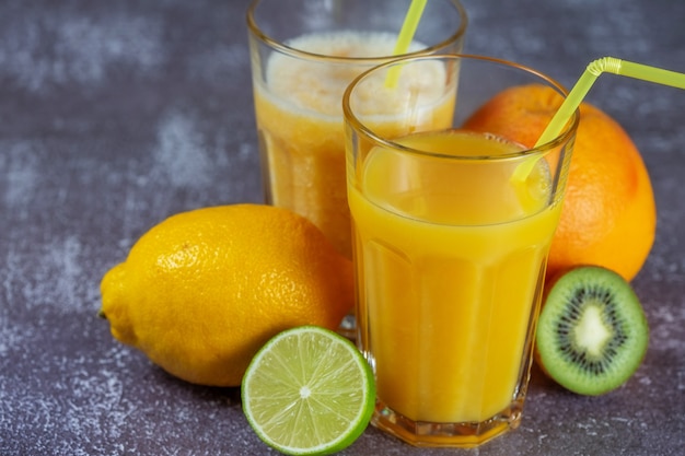 Freshly squeezed orange juice and banana and orange smoothies
in tall glasses with a straw surrounded by fruits on a gray
concrete background. the concept of slimming, bring the figure into
shape.