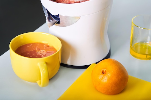 Photo freshly squeezed orange juice in yellow cup fresh cut orange and juicer on kitchen