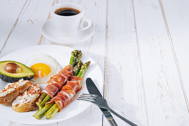 Fried asparagus wrapped in jamon fried eggs bruschetta with soft cheese and avocado on a white plate and a cup of freshly brewed coffee