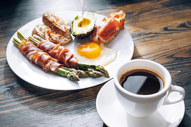 Fried asparagus wrapped in jamon fried eggs bruschetta with soft cheese and avocado on a white plate and a cup of freshly brewed coffee