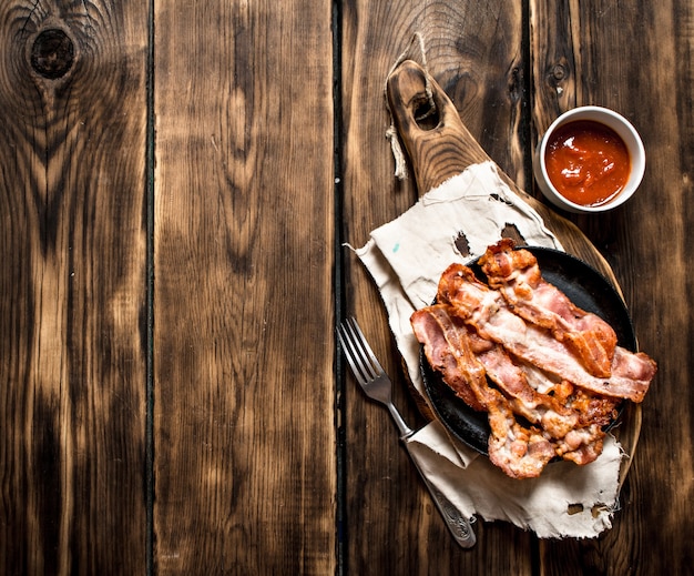 Fried bacon in a frying pan and tomato sauce. On a wooden table.