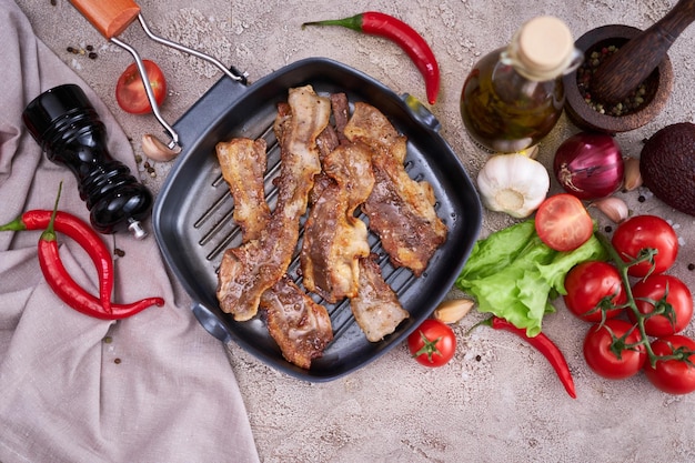 Fried bacon pieces in a skillet and vegetables at domestic kitchen stone or concrete table