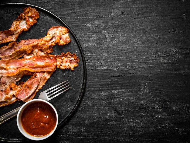 Fried bacon with sauce and greens. On a black wooden background.