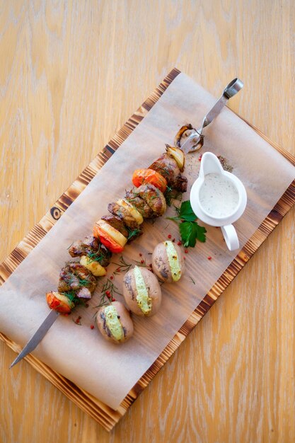 Fried barbeque meat with vegetables on a skewer and potatoes\
grilled bbq decorated with vegetables and sauce on wooden table and\
parchment paper