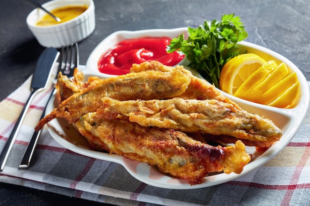 Fried in batter capelins with tomato sauce, sliced lemon on a plate on a concrete table with fork and knife, horizontal view from above, close-up