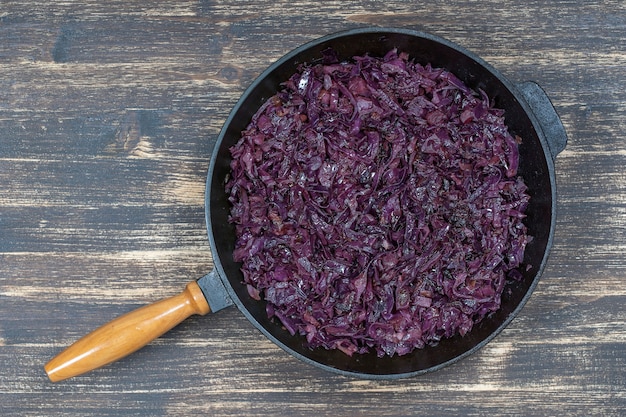 Fried blue cabbage with onion, apple and garlic in frying pan on wooden table. Top view, close up