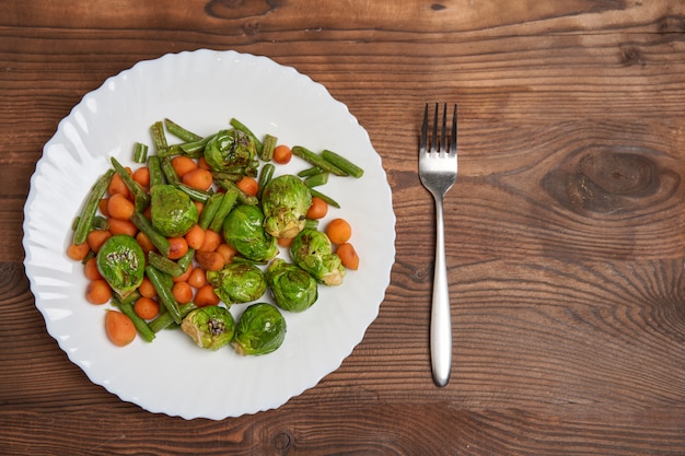 Fried Brussels sprouts carrots and green beans lie on a white plate that stands on a wooden table.