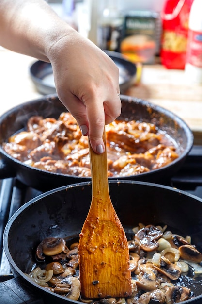 Fried champignons with onions in a frying pan with a wooden\
spatula stirs a female hand