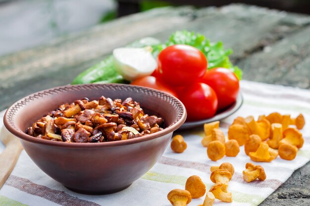 Fried chanterelles with onion in rustic bowl and plate with fresh vegetables for salad on surface