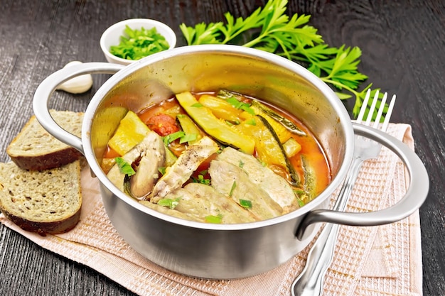 Fried chicken breast slices with stewed zucchini, tomatoes and garlic in a saucepan on napkin, bread, parsley and a fork on wooden board background