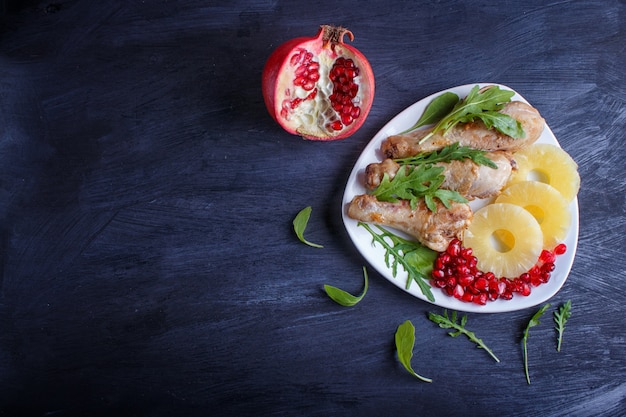 Fried chicken legs with arugula, pineapple and pomegranate seeds on black wooden background.