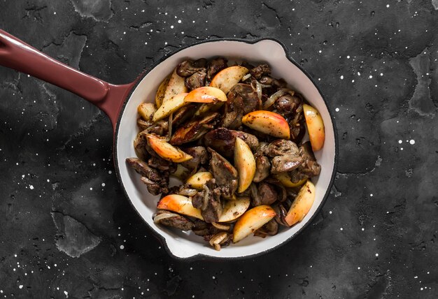 Photo fried chicken liver with onions and apples in a ceramic frying pan on a dark background top view