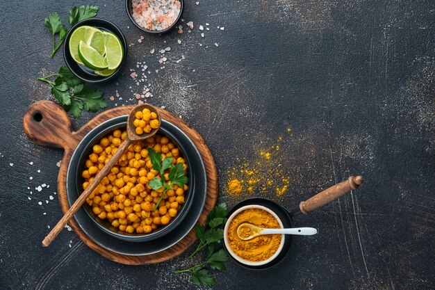 Fried chickpeas with turmeric with parsley and lime in black plate on an old black table background. Roasted spicy chickpeas or Indian chana or chole, popular snack recipe. Top view.